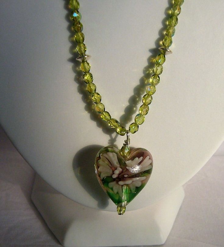 Ref: P016.     Green glass heart with a lilac flower within the glass measuring 1 inch across.  The wire has small sparkly green beads and silver plated spacers.  Length is 20 inches and has a fancy silver plated heart clasp. Green Heart-shaped Beaded Necklace Gift, Handmade Green Jewelry For Valentine's Day, Green Glass Jewelry With Faceted Beads, Green Wire Wrapped Czech Glass Beaded Necklaces, Wire Wrapped Green Glass Jewelry, Green Wire Wrapped Glass Jewelry, Green Wire Wrapped Beaded Necklace With Czech Glass, Green Heart-shaped Beaded Necklace, Green Glass Flower-shaped Jewelry