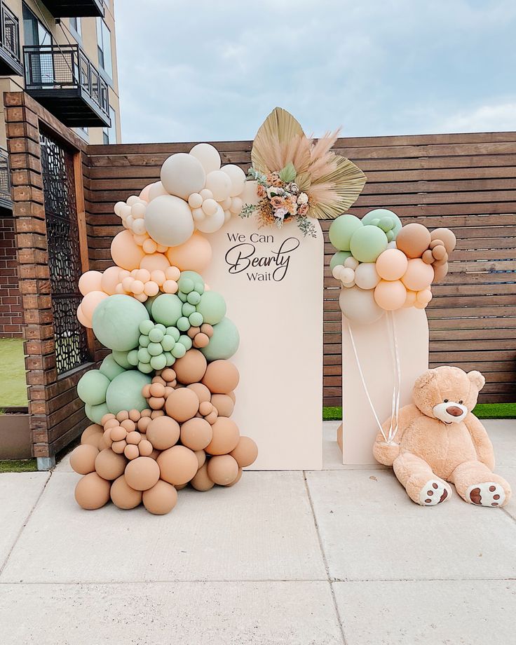 a teddy bear sitting in front of a sign with balloons attached to it's sides