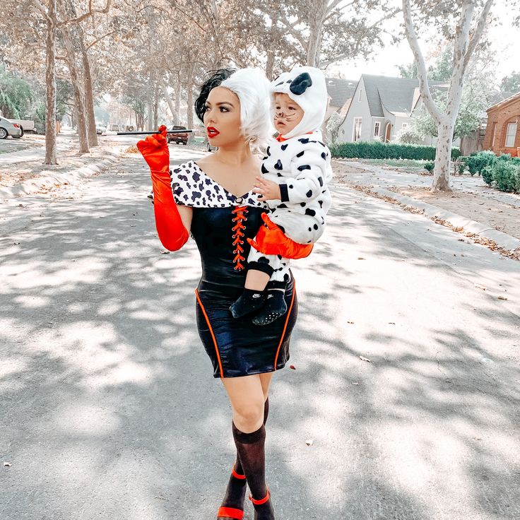 a woman holding a baby in her arms while walking down a street with trees and houses behind her