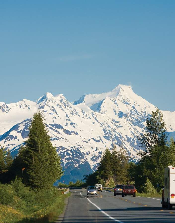 the mountains are covered in snow as cars drive down the road with trees on both sides