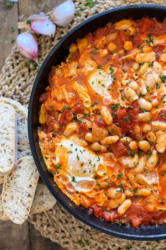 a skillet filled with chickpeas and tomato sauce next to garlic bread on a mat