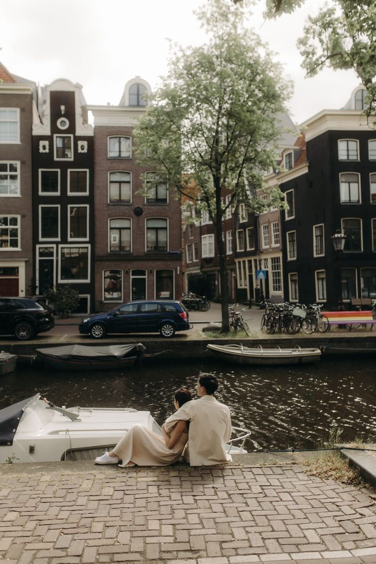 a man and woman sitting on the edge of a river in front of some buildings