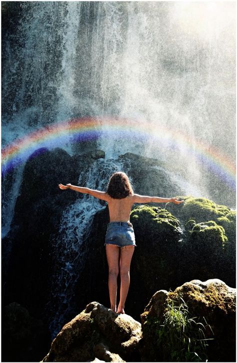 a woman standing in front of a waterfall with her arms wide open and rainbow coming out