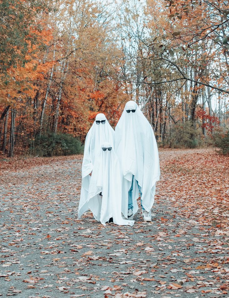 two people dressed in white ghost costumes walking through the woods with leaves on the ground