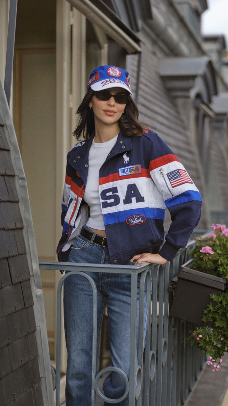 a woman standing on a balcony with her hands in her pockets and wearing a baseball cap