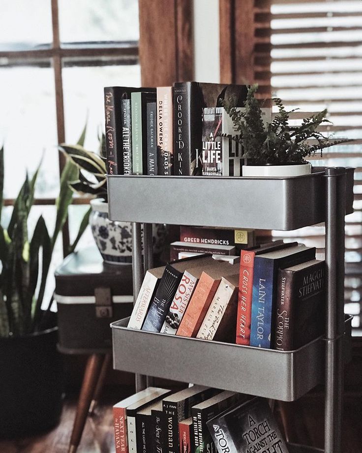 there is a metal shelf with books on it in front of a window and potted plants