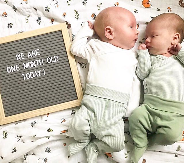 two babies laying next to each other on a bed with a chalkboard that says we are one month old today