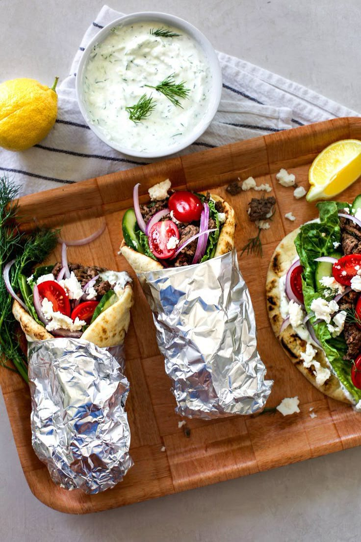 two pita sandwiches on a wooden platter with silver foil wrappers and fresh vegetables