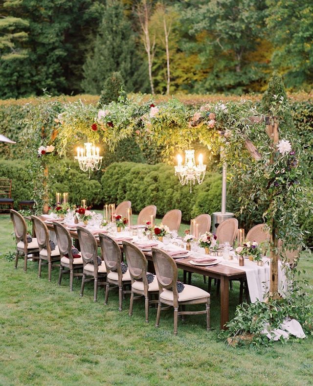 an outdoor dinner table set up with chandeliers and flowers in the center, surrounded by greenery