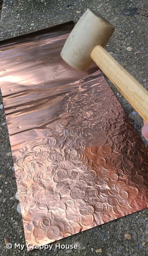 a person using a hammer to cut metal sheet with copper pattered on it and another piece of wood sticking out of the ground