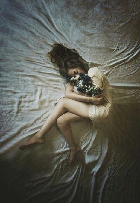 a woman laying on top of a bed covered in white sheets holding a bouquet of flowers