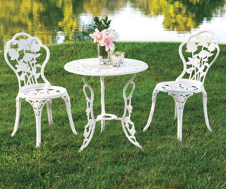 two white chairs and a table in the grass near water with flowers on top of it