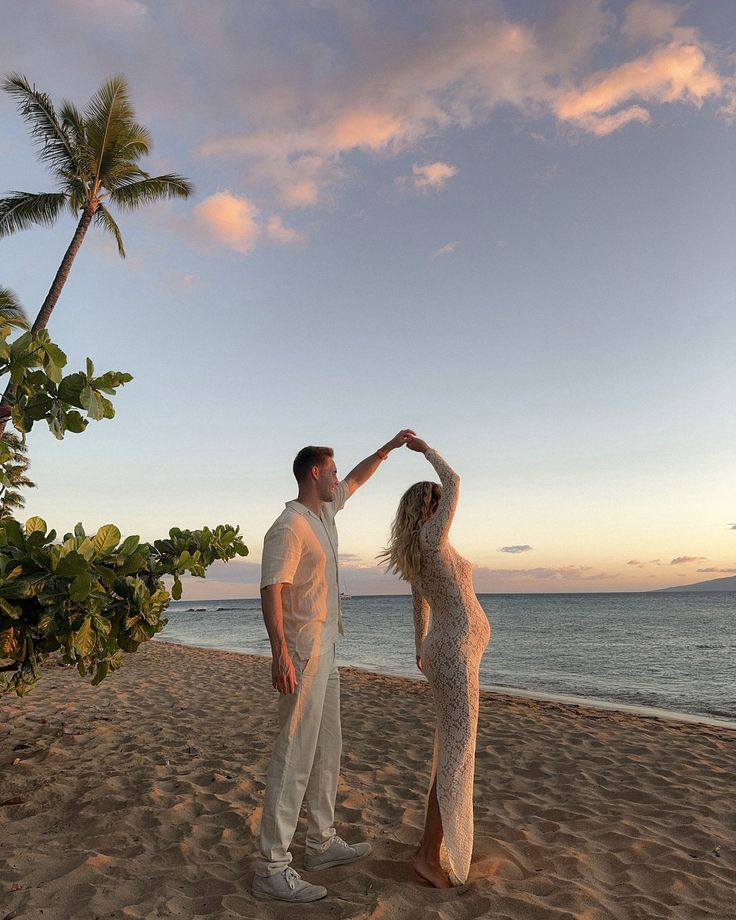 a man and woman standing on top of a sandy beach next to the ocean at sunset