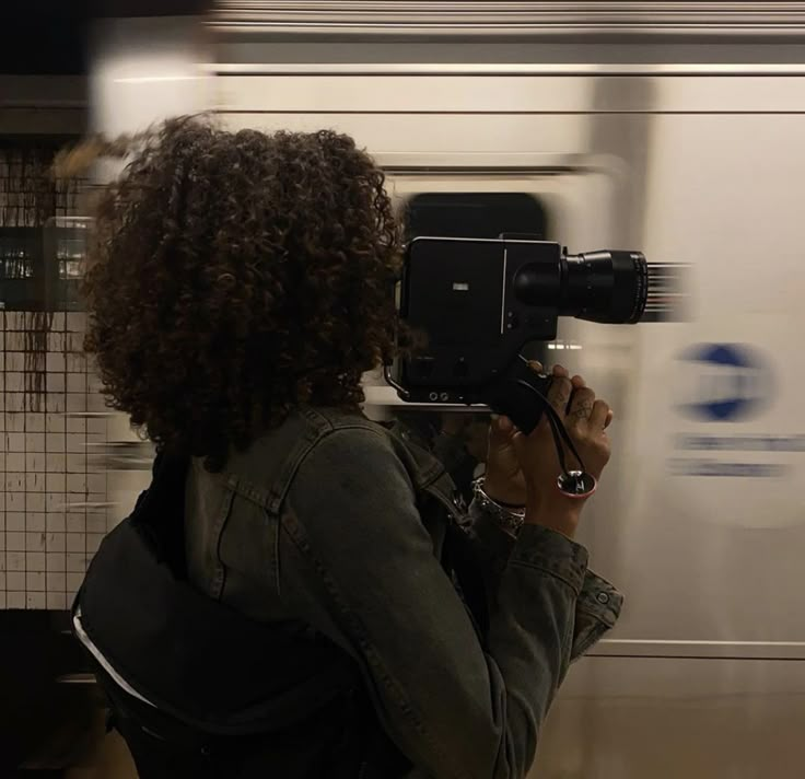 a woman with curly hair is holding a camera in front of a moving subway train