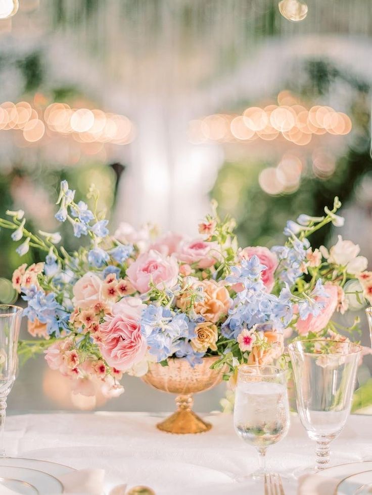 an arrangement of flowers in a vase on a table with wine glasses and silverware