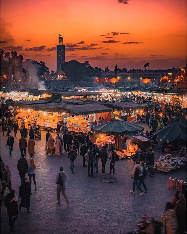 an outdoor market with people walking around it at sunset or dawn in the background is a clock tower