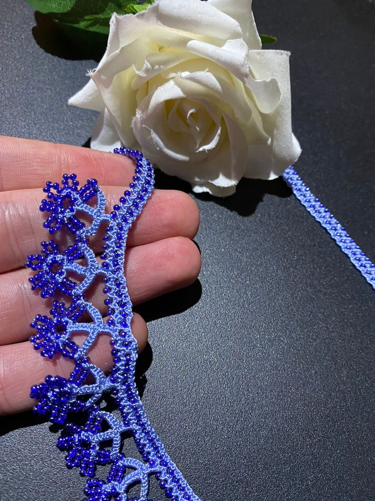 a hand holding a blue beaded necklace next to a white rose on a table