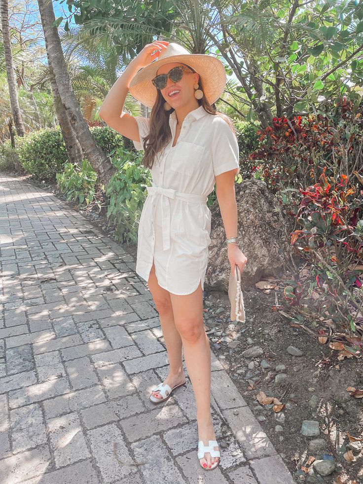 a woman in a white dress and hat walking down a brick path with palm trees behind her