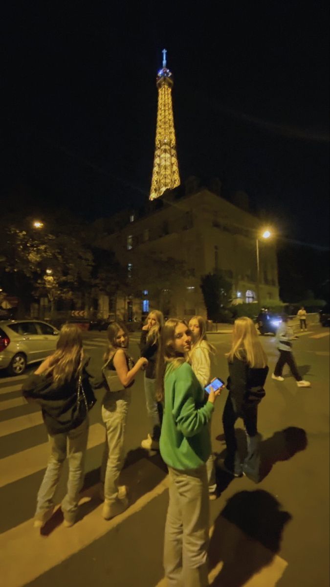 group of people standing in front of the eiffel tower at night, looking at their cell phones