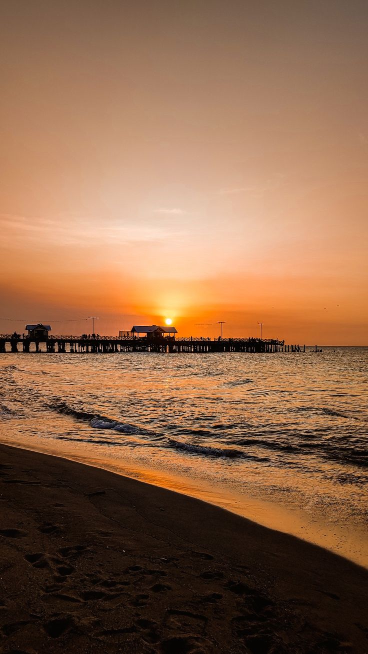 the sun is setting over the ocean with a pier in the distance