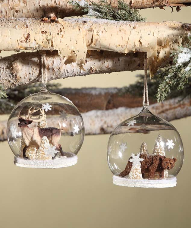 two glass ornaments hanging from a tree branch with snow and trees in the background,