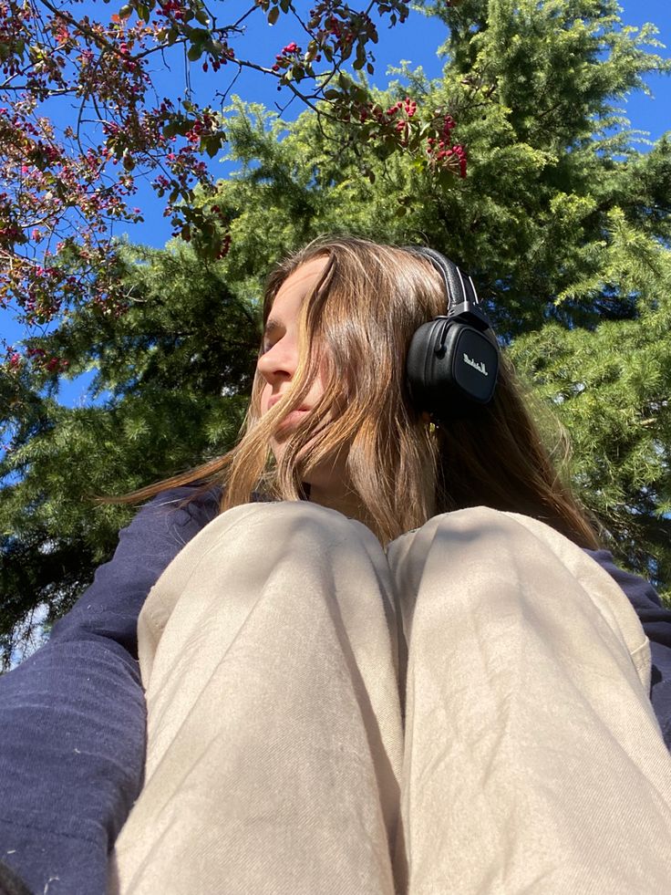 a woman with headphones on sitting in front of trees and looking up at the sky