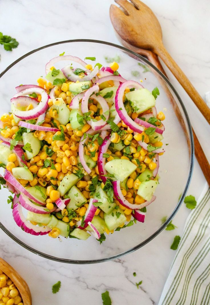 a glass bowl filled with corn and cucumber salad next to wooden spoons