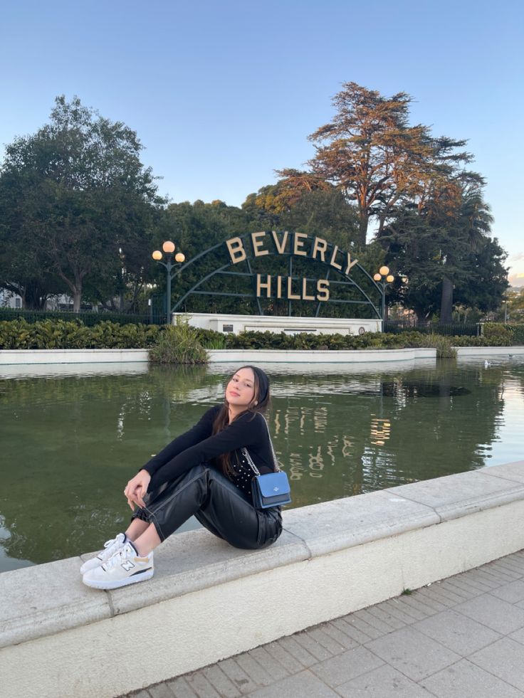 a woman is sitting on the edge of a pond in front of beverly hills sign