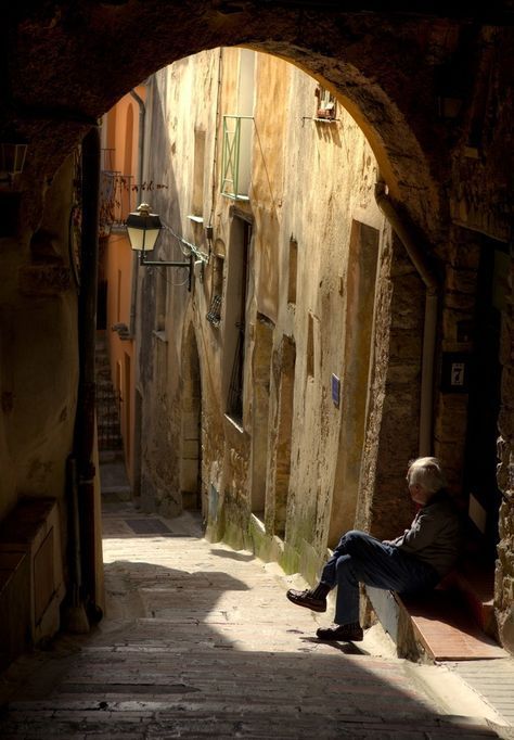 an old man sitting on a bench in the middle of a narrow alleyway between two buildings