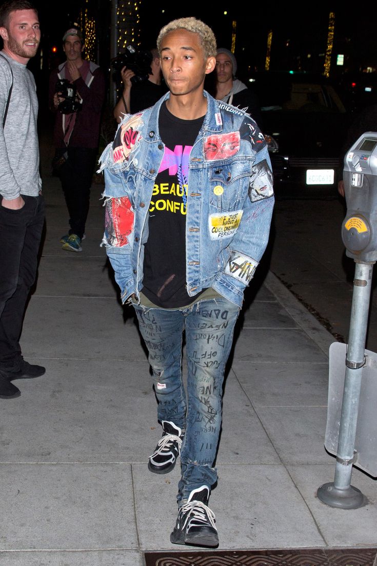 a young man wearing ripped jeans and a denim jacket with patches on it is walking down the street