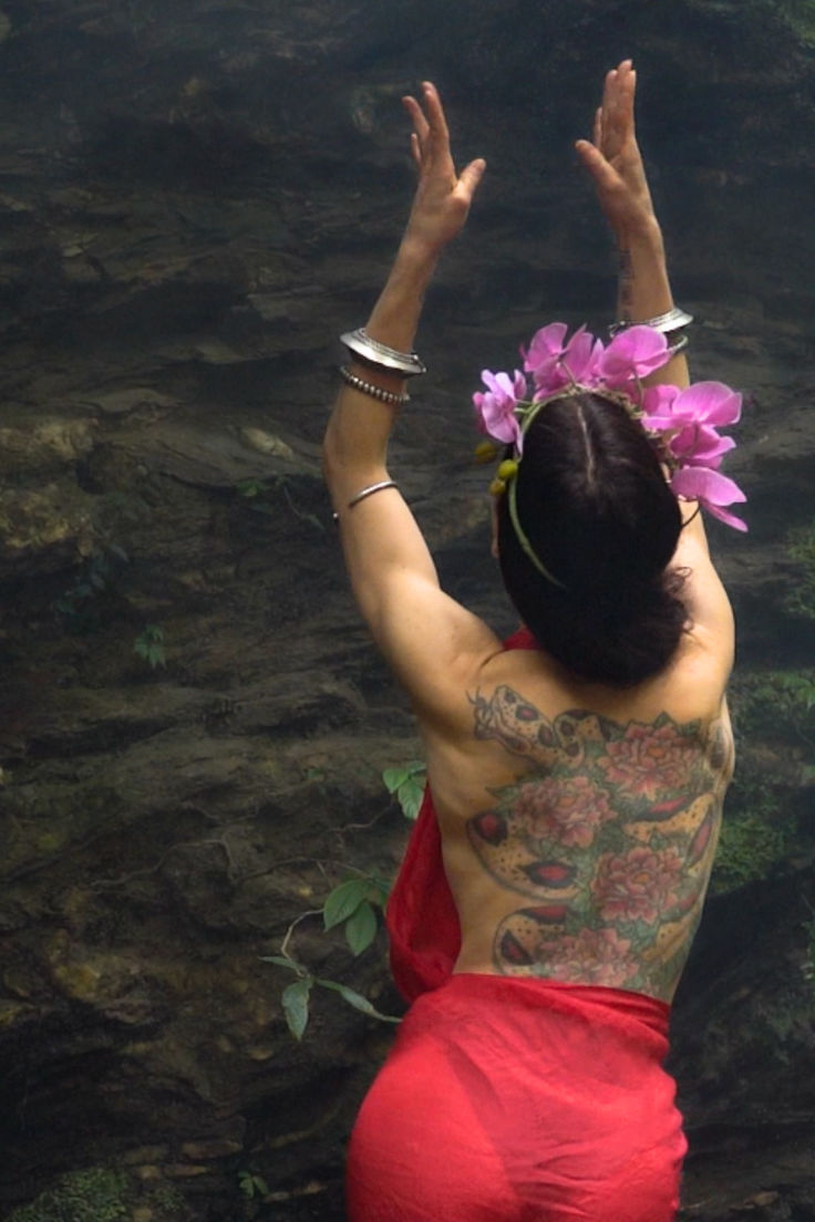 a woman in a red skirt standing next to a body of water with her hands up