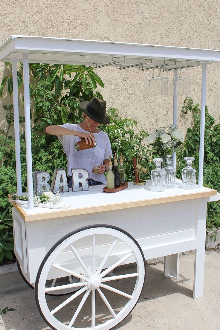 a man standing behind a white cart filled with bottles and glasses on top of it