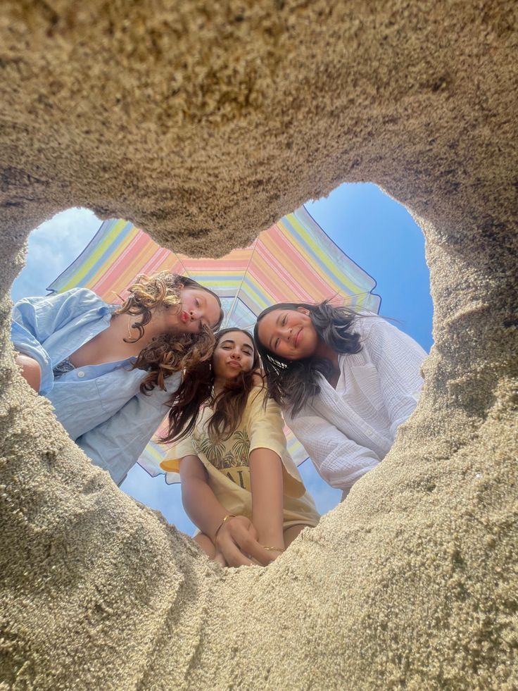 three girls are standing in the sand with an umbrella over their head and looking at the camera