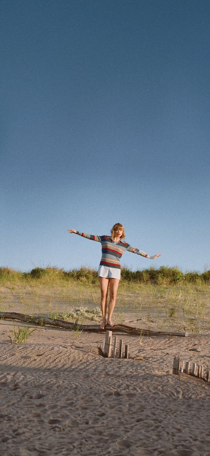 a woman in striped shirt and white shorts standing on sand with arms spread wide open