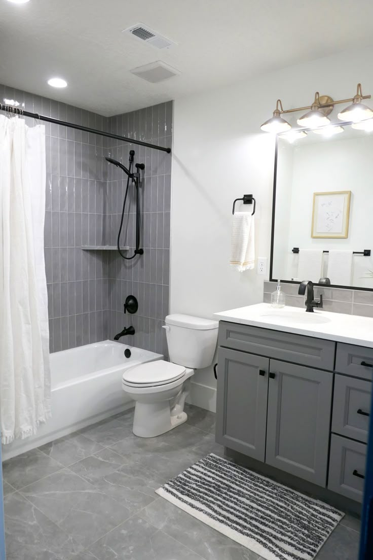 A bathroom with a tub/shower combo that has tub to ceiling gray glass subway tile installed vertically. The toilet is between the tub and single-sink vanity, which has darker gray cabinets and white Quartz countertops. The floor has large rectangular tiles that are gray with white veins. White Bathroom With Grey Floor, Grey Bathroom Styling, Modern Bathroom Grey And White, White And Dark Grey Bathroom, Gray Toilet Bathroom, Gray Sink Bathroom, Small Bathroom White And Grey, Small Bathroom With Grey Vanity, Bathroom Interior Grey And White