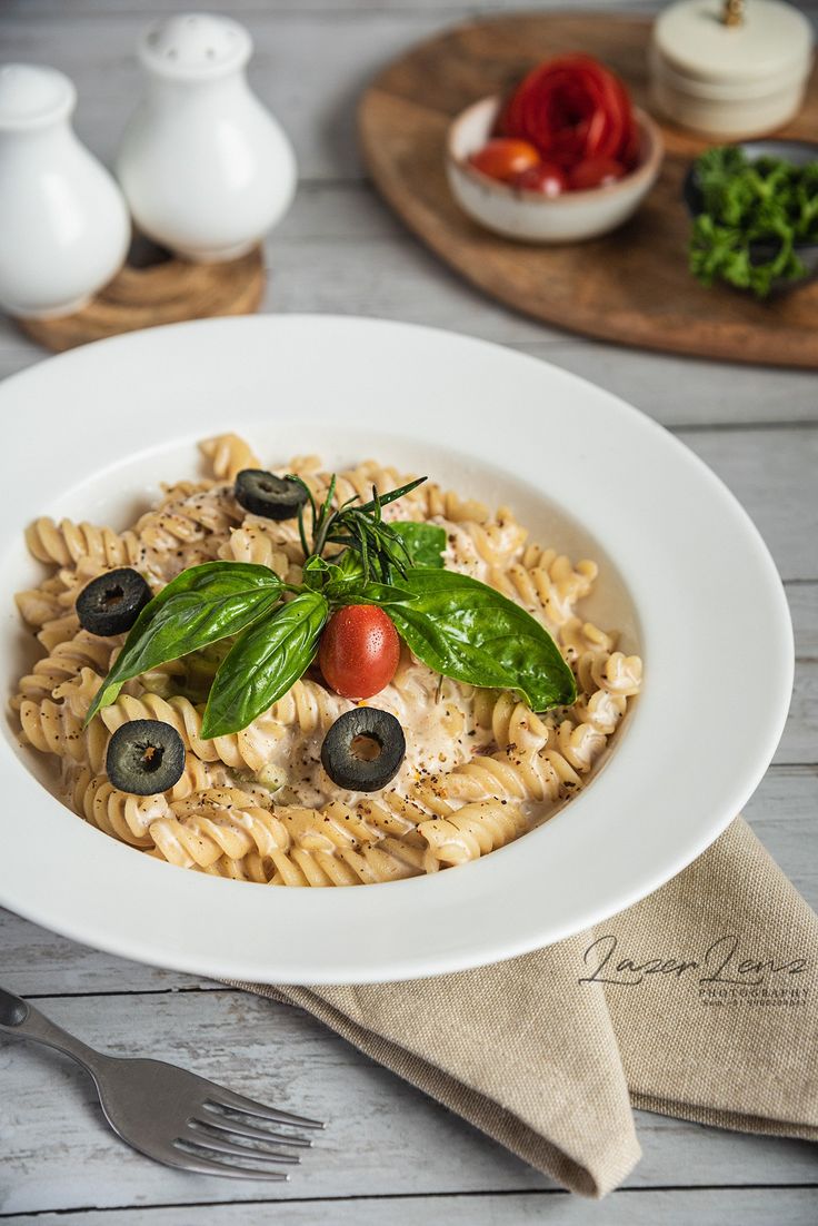 a white bowl filled with pasta and olives on top of a wooden table next to silverware