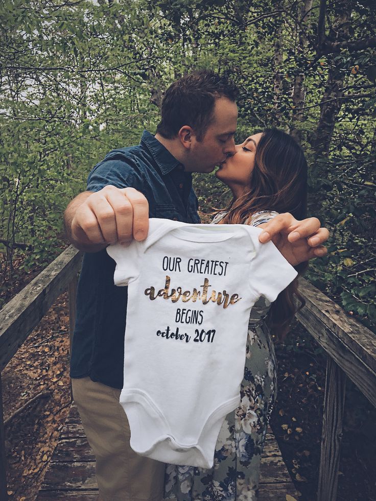 a man and woman kissing while holding up a baby's bodysuit with the words our greatest adventure begins on it