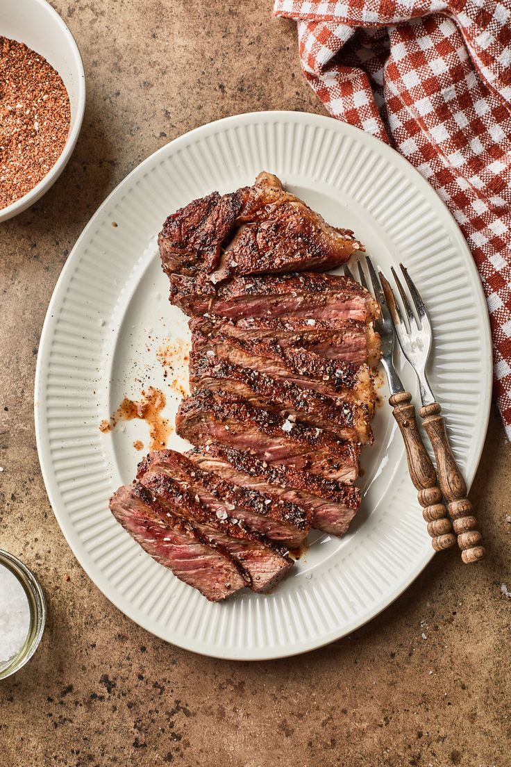 a plate with steak on it next to a bowl of seasoning and a fork