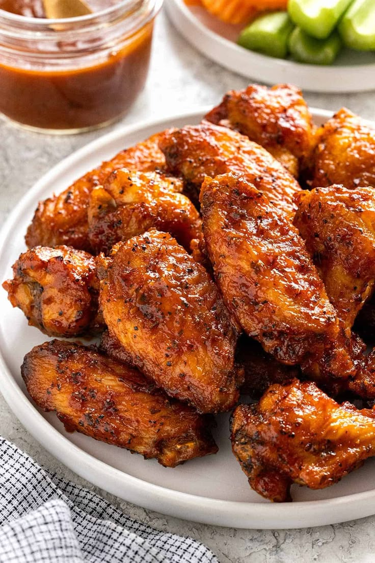 some chicken wings are on a white plate with pickles and carrots in the background