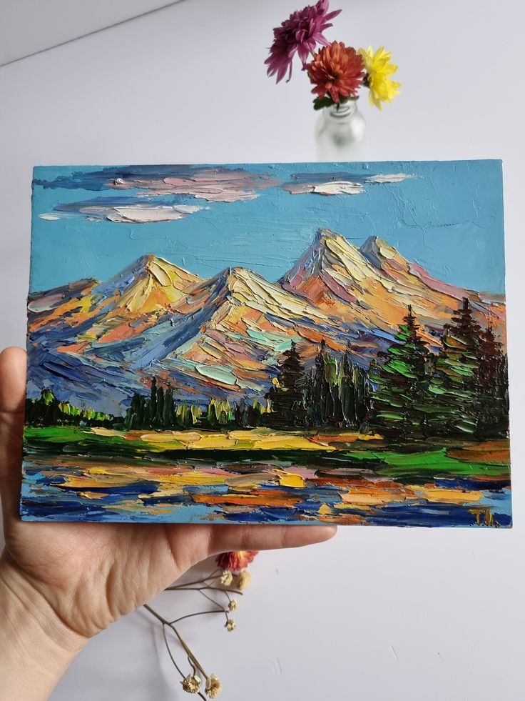 a hand holding up a piece of art with mountains in the background and trees on it