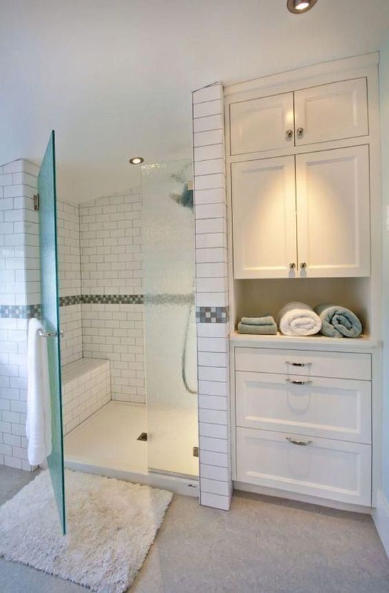 a bathroom with white tile and glass shower door, rugs on the floor and cabinets