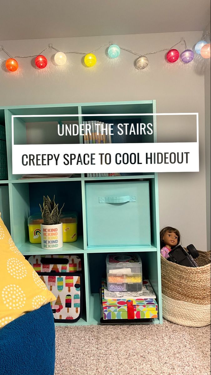 An aqua colored bookshelf sits in an under the stairs area with colored string lights and a basket of kid toys. Under The Stairs Toy Storage, Under The Stairs Library, Understairs Kids Space, Understairs Kids Space Play Areas, Under Stairs Closet Playroom, Under Stairs Toy Storage, Under Stairs Ideas For Kids, Under Stairs Nook Kids, Kids Under Stairs Play Areas