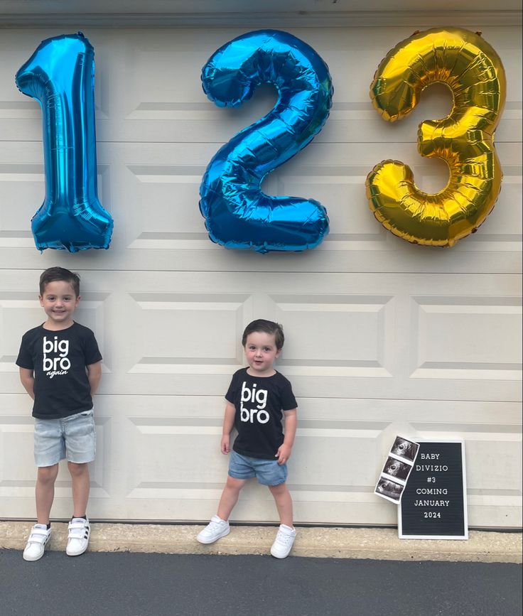 two young boys standing next to each other in front of a garage door with blue and gold balloons