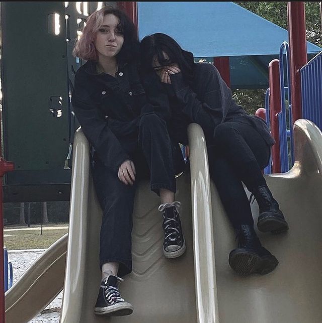 two women sitting on top of a slide in a playground area, one with pink hair and the other with black shoes