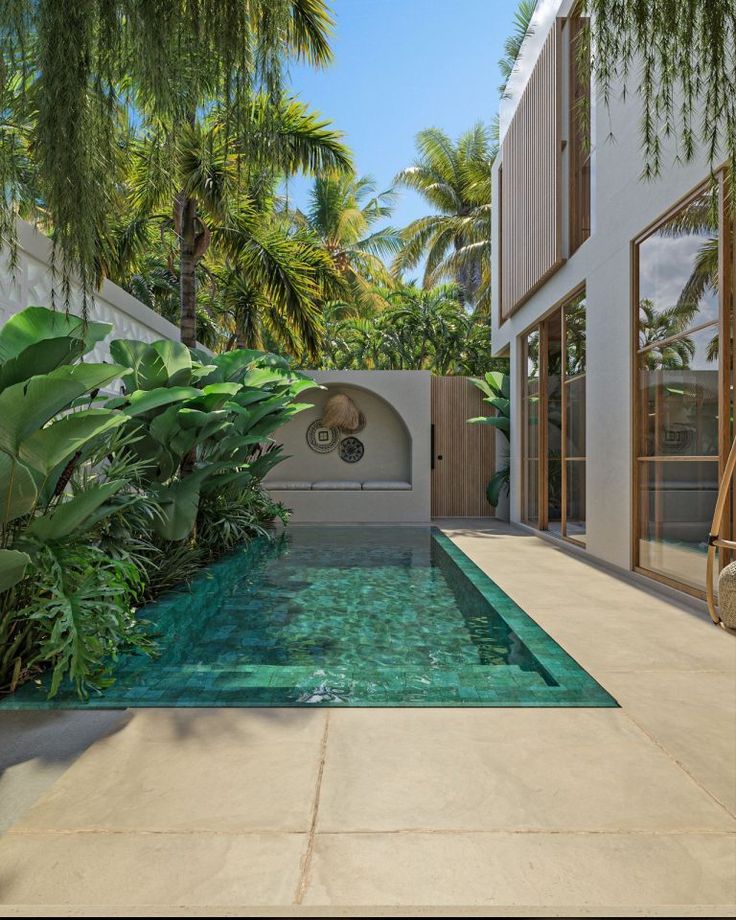 an outdoor swimming pool surrounded by palm trees and greenery in front of a house