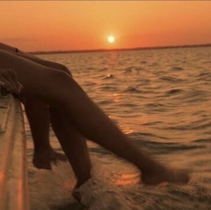 a woman is sitting on the edge of a boat at sunset, with her legs in the water