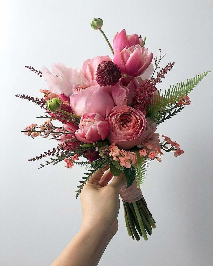 a hand holding a bouquet of pink flowers and greenery in front of a white wall