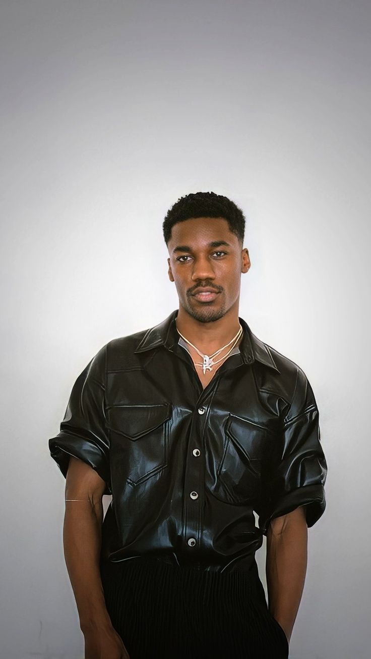 a man wearing a leather shirt and black pants standing in front of a white wall