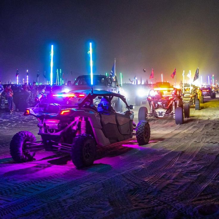 several vehicles are parked in the dirt at night time with bright lights on them and people standing around