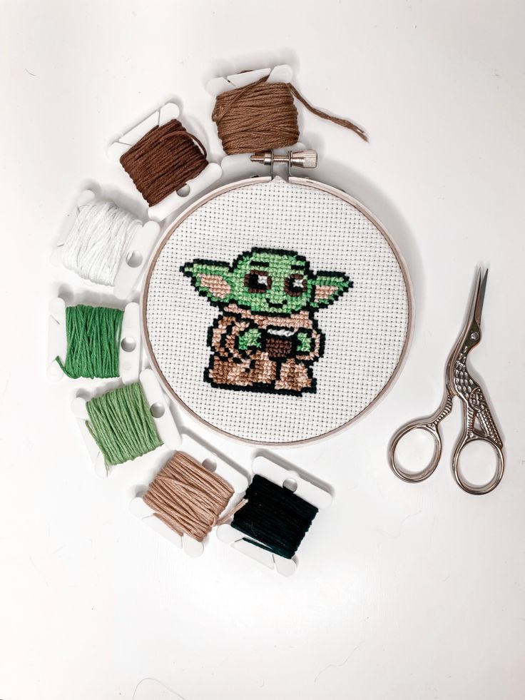 a cross stitch kit with yarn, scissors and thread on a white table next to it is an image of the baby yoda from star wars